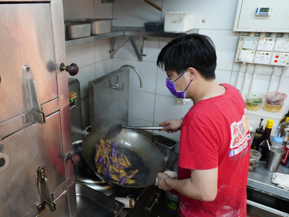廚房裡，一位年輕大廚正俐落地將茄子放入熱油下鍋，拋著大鐵鍋猛火翻炒。