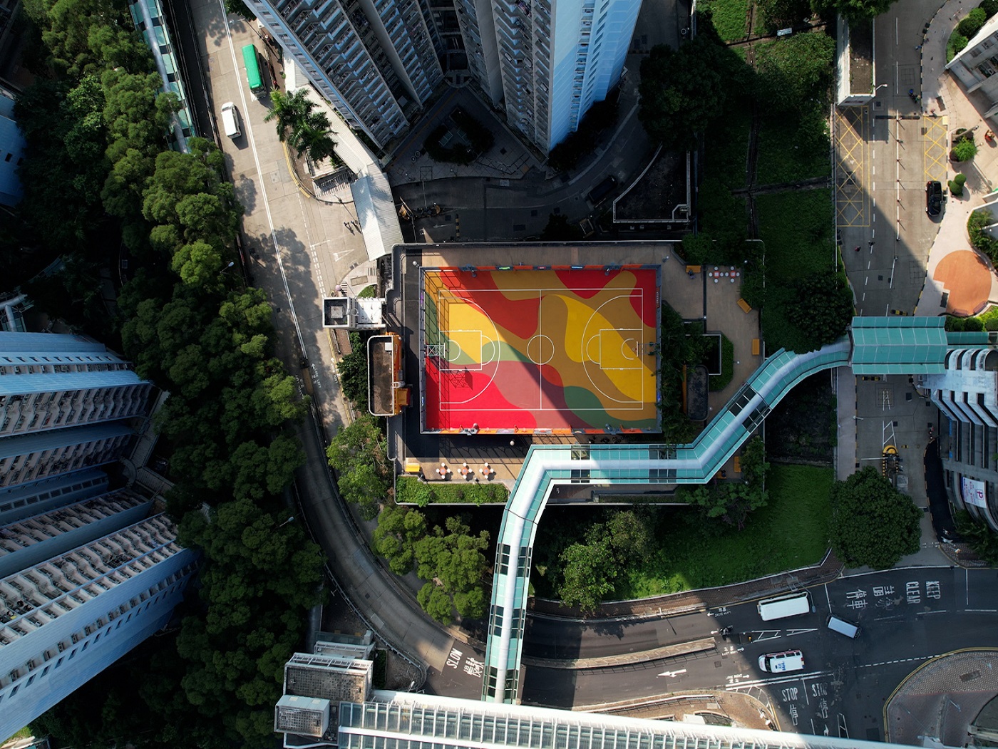 Seen from above, the rooftop basketball court bursts with bold red, orange, and yellow tones. Its dynamic camo-style flooring radiates youthfulness and vitality.